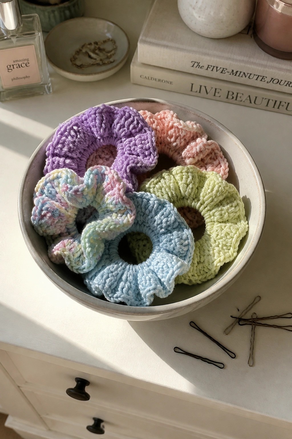 Colorful crocheted scrunchies fill a ceramic bowl on a sunlit white vanity with books.