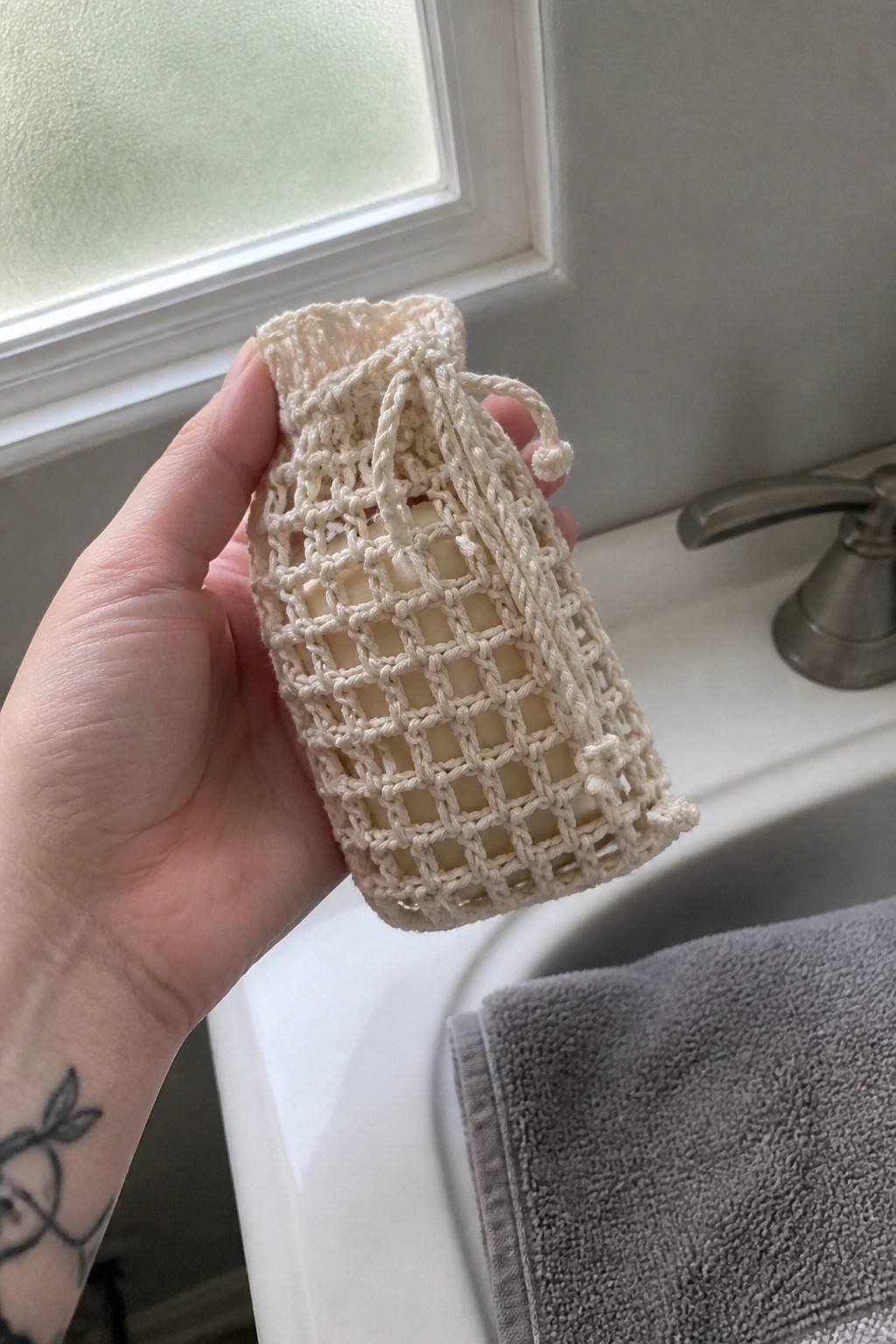 Hand holding soap bar in cream crocheted mesh pouch above bathroom sink