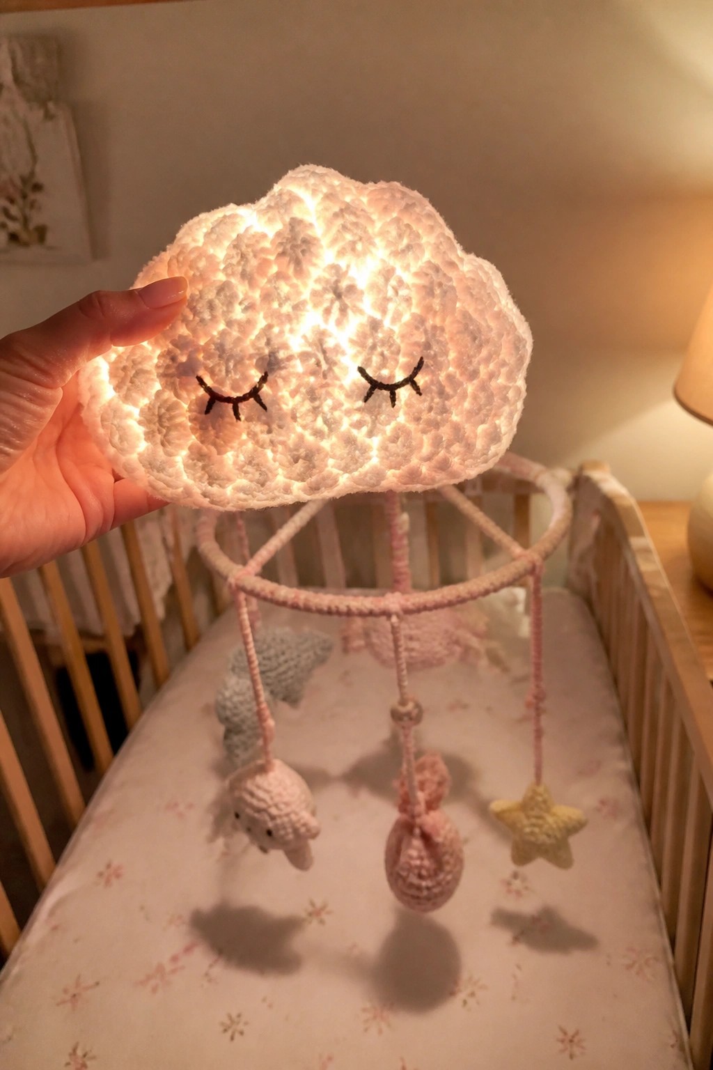 Hand holding glowing crocheted cloud with closed eyes above pastel crib mobile.