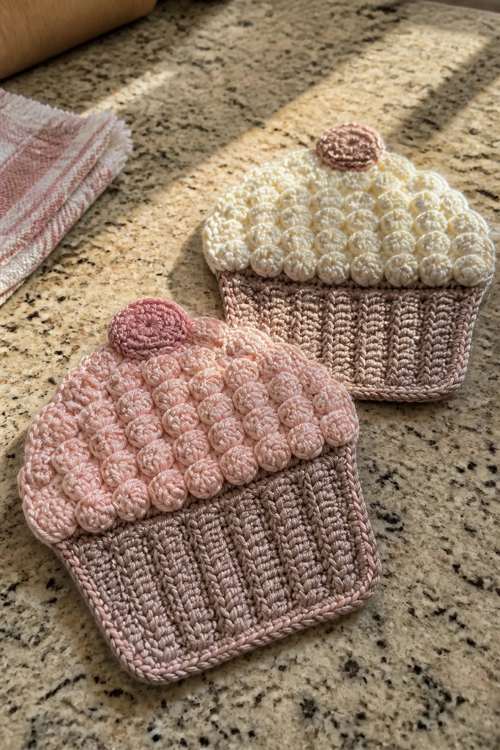 Two crocheted cupcake-shaped coasters sit on a granite countertop next to a folded cloth.