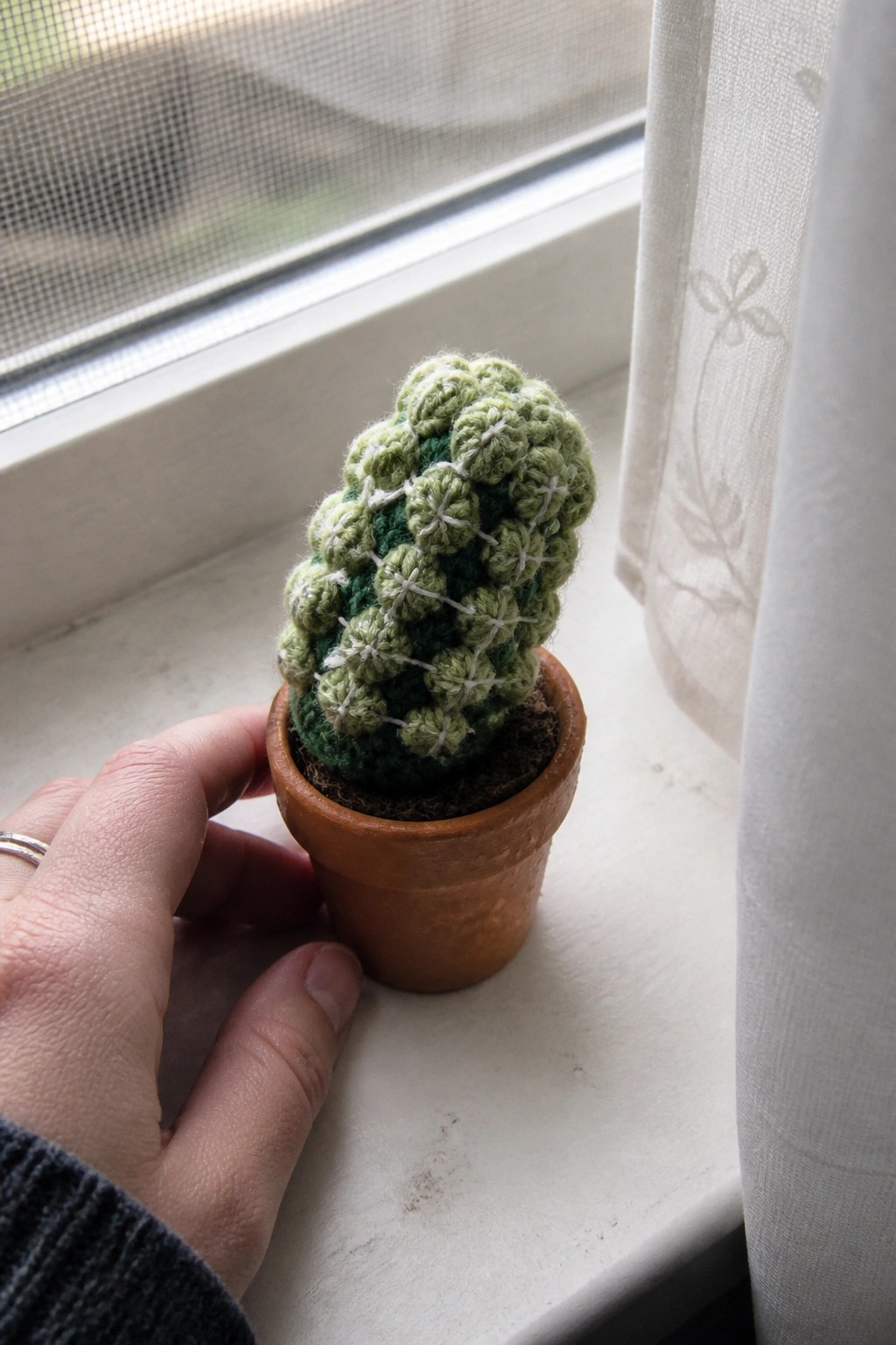 Hand holding crocheted green cactus in terracotta pot on white windowsill with curtain