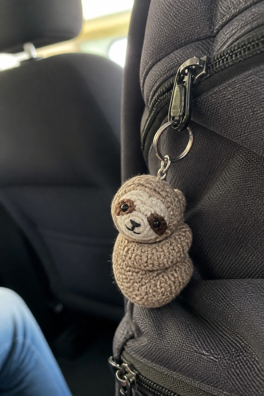 Crocheted sloth keychain attached to zipper of gray backpack in car