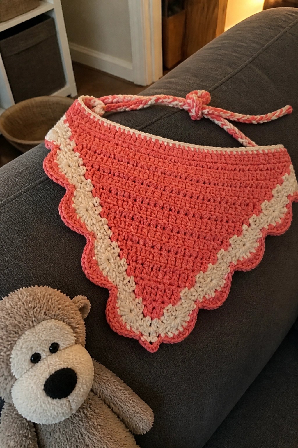 Pink crocheted scalloped bib with ties beside teddy bear on gray couch