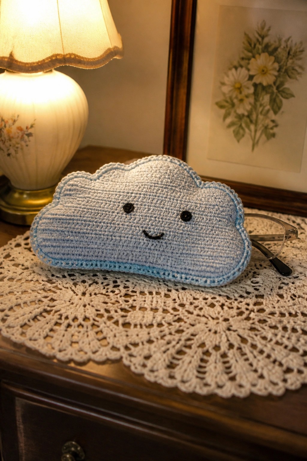 Light blue crocheted cloud with smiley face on lace doily beside lamp and glasses