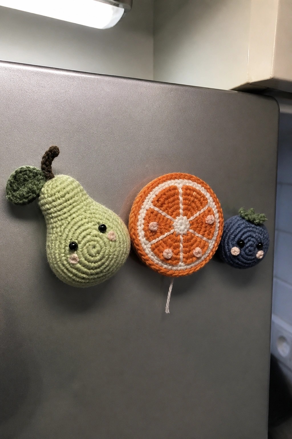 Three crocheted fruit magnets on a gray refrigerator: a green pear with stem and leaf, a flat orange slice with white stitching, and a small blue berry with green top.