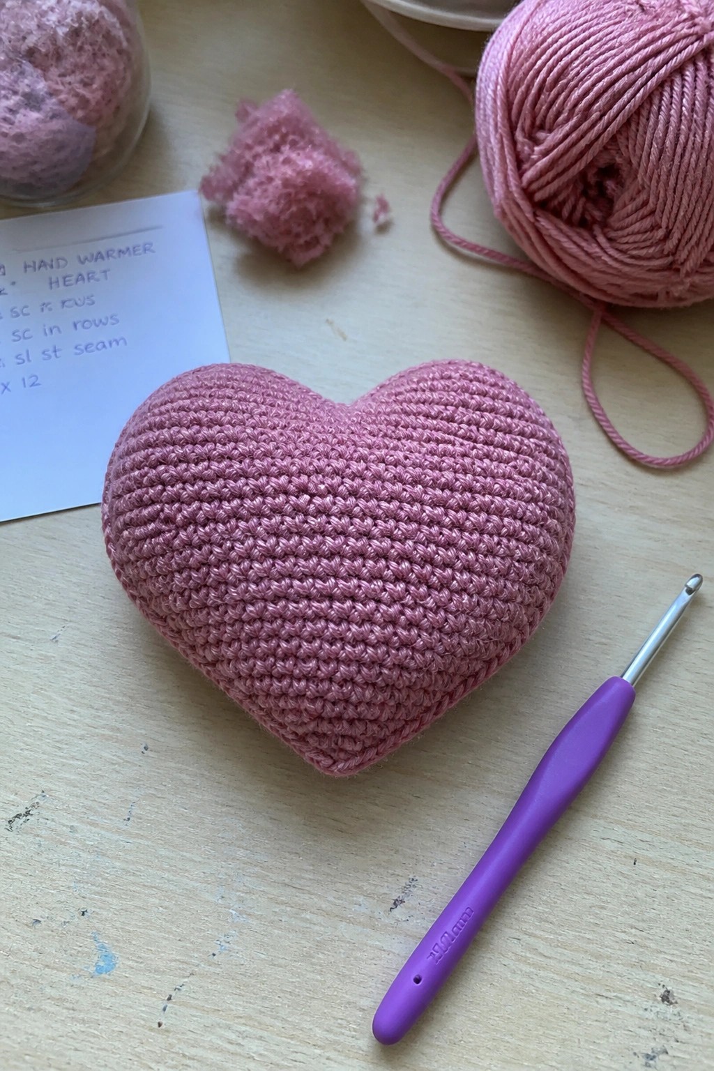 Pink crocheted heart hand warmer with purple hook, yarn, and notes on table.
