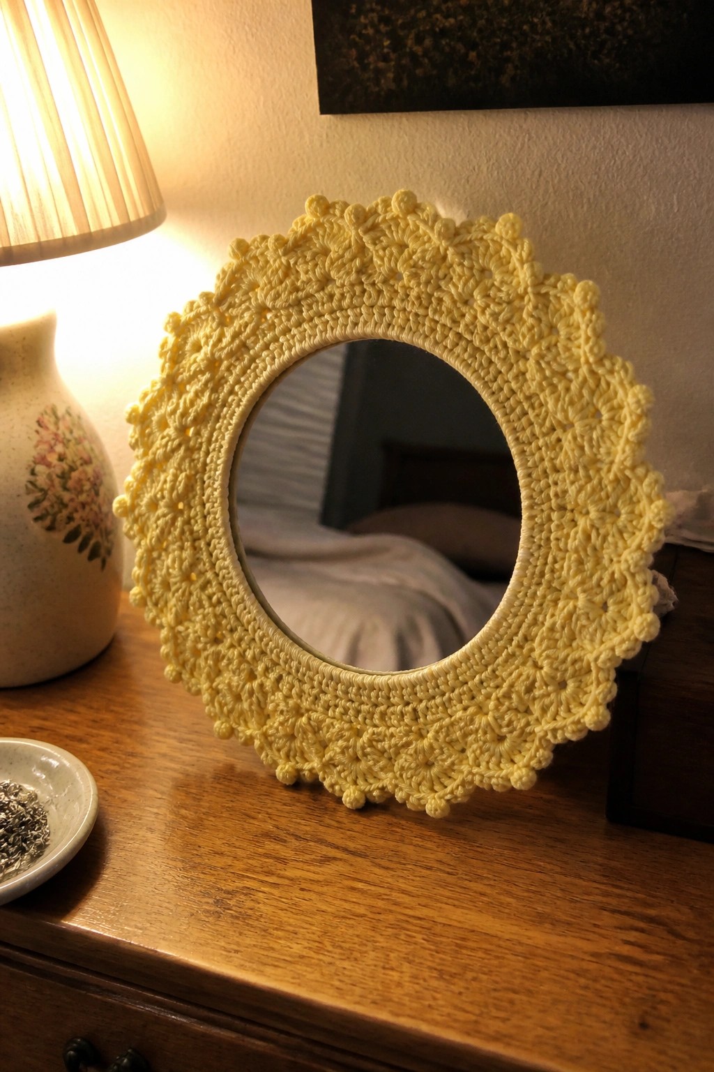 Yellow crocheted round mirror on wooden dresser beside lamp and floral vase
