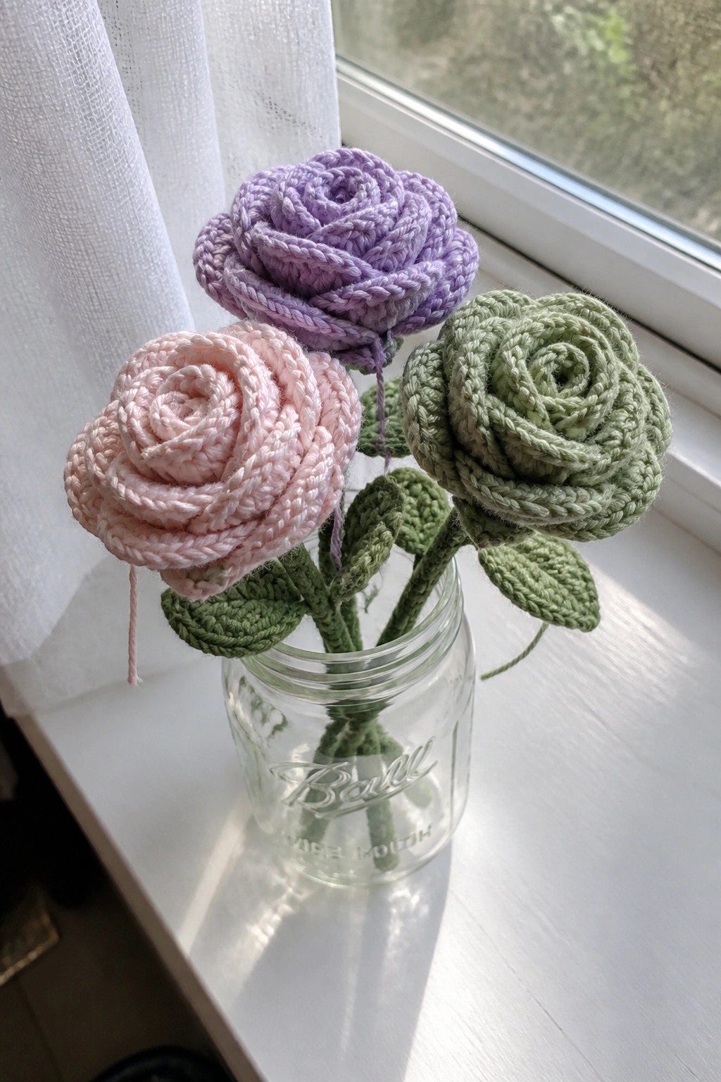 Three crocheted roses in pink, purple, and green inside a glass jar on a windowsill.