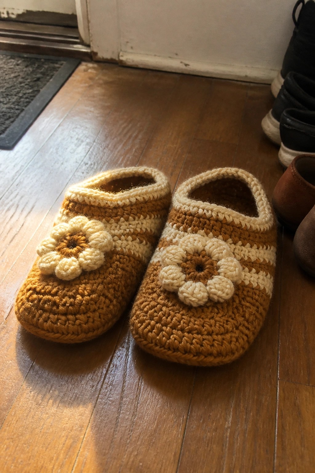 Pair of brown and cream crocheted slippers with flower accents on wooden floor.