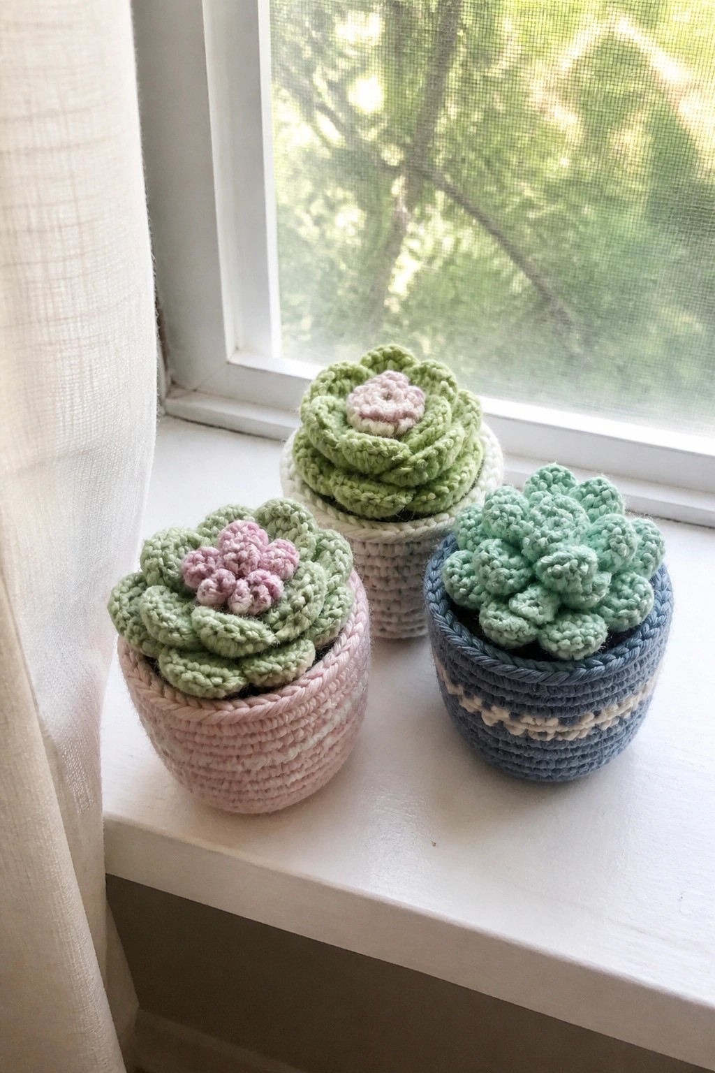 Three crocheted succulents in pink, white, and blue pots sit on a white windowsill.