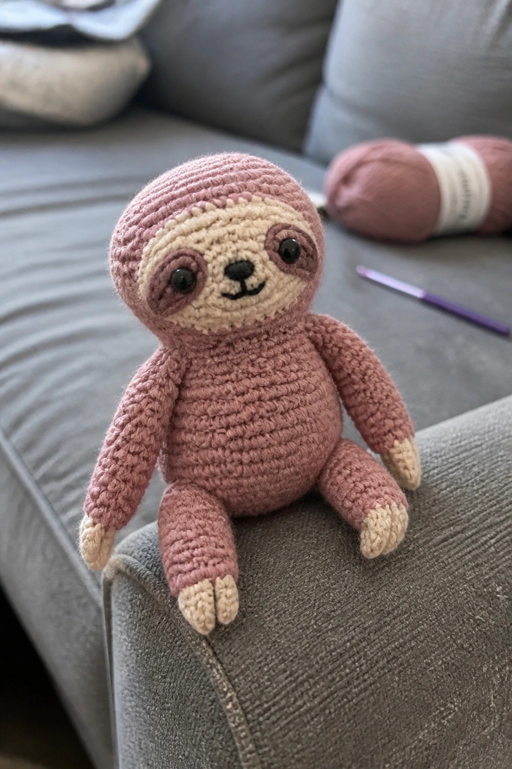Pink crocheted sloth toy on gray couch with yarn ball and hook nearby.
