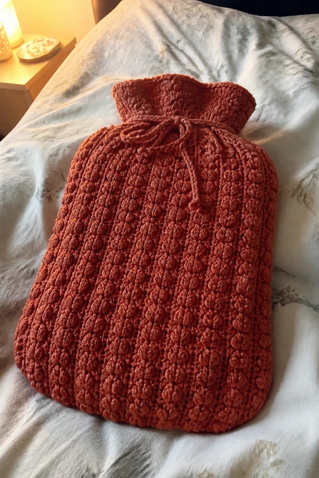 Reddish-orange knitted hot water bottle cover on rumpled white bedsheets