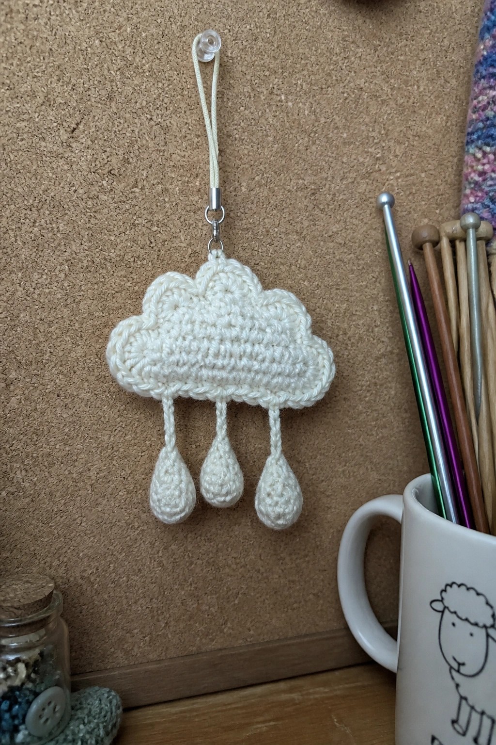Crocheted white cloud with three dangling raindrops beside knitting needles in sheep mug