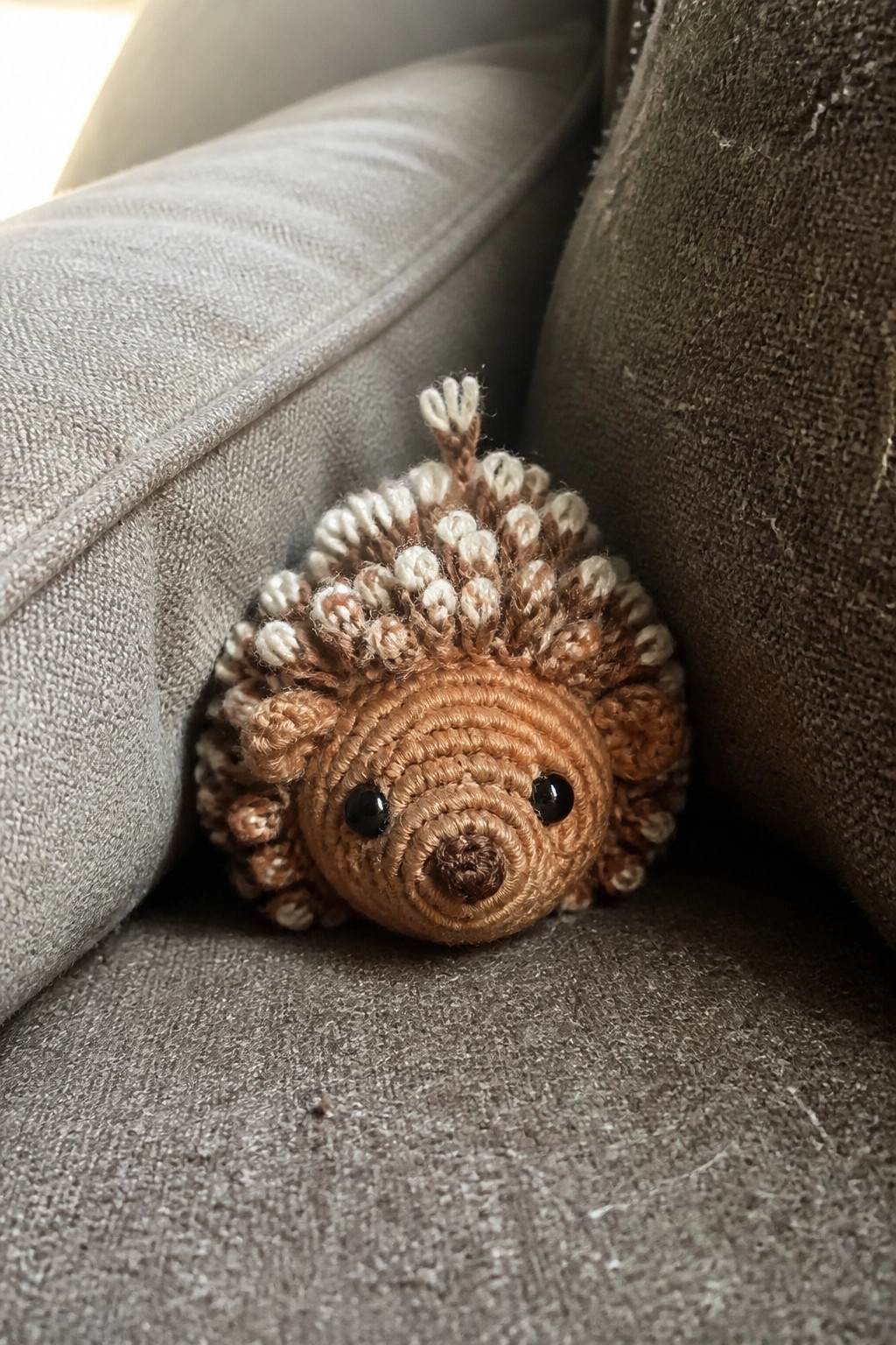 Small brown crocheted hedgehog toy with white spines nestled in gray couch cushions.