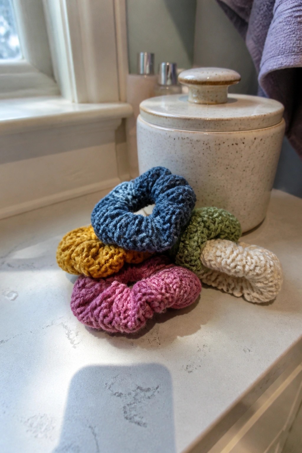 Pile of multicolored crocheted scrunchies beside a ceramic jar on a bathroom counter.