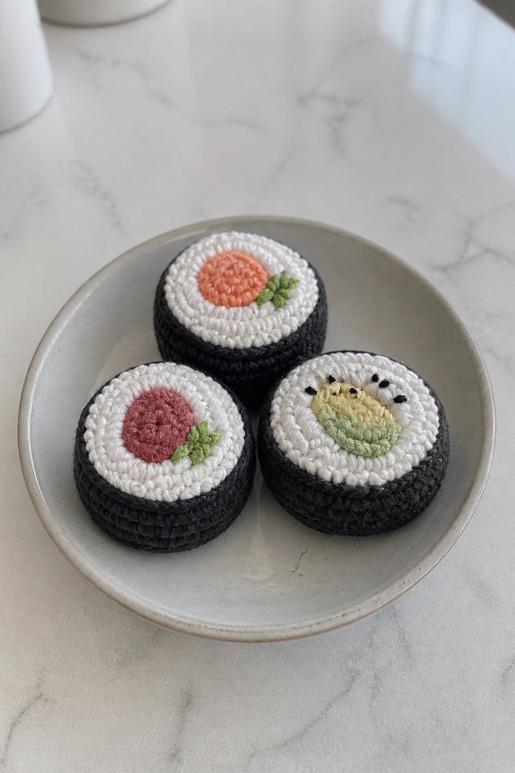 Three crocheted sushi rolls with pink, orange, and avocado centers on a plate.