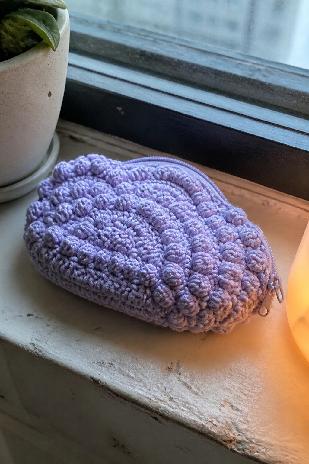 Purple crocheted zippered pouch on windowsill beside potted plant and glowing lamp