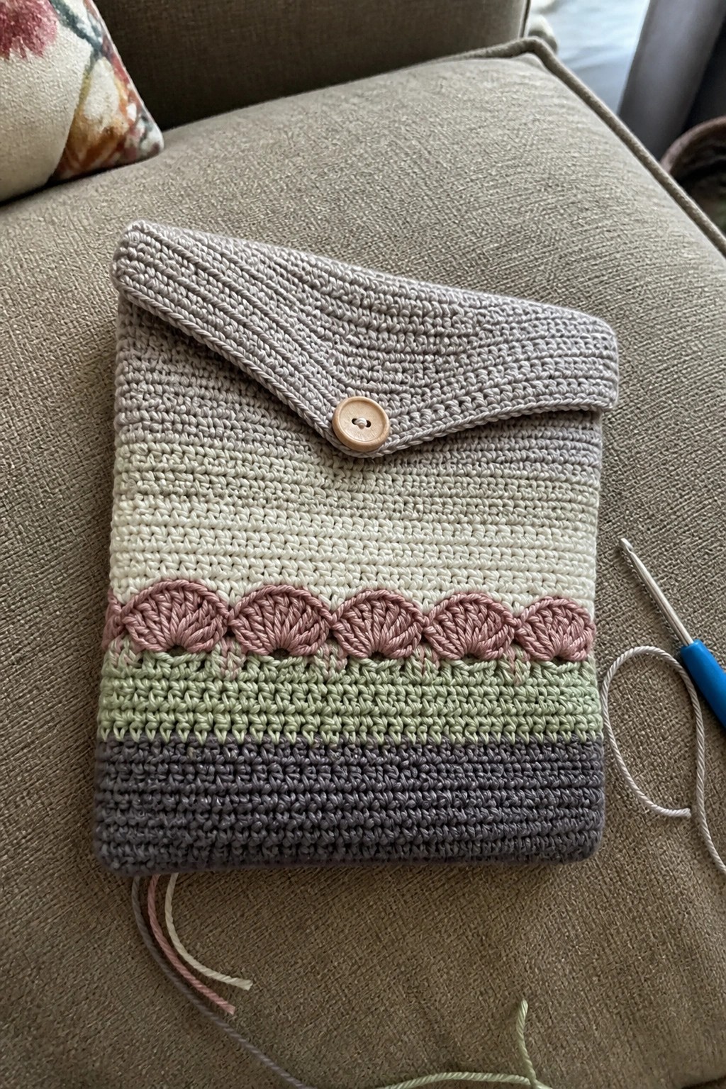 Crocheted envelope pouch with striped bands, pink scallops, beige flap, and button on couch