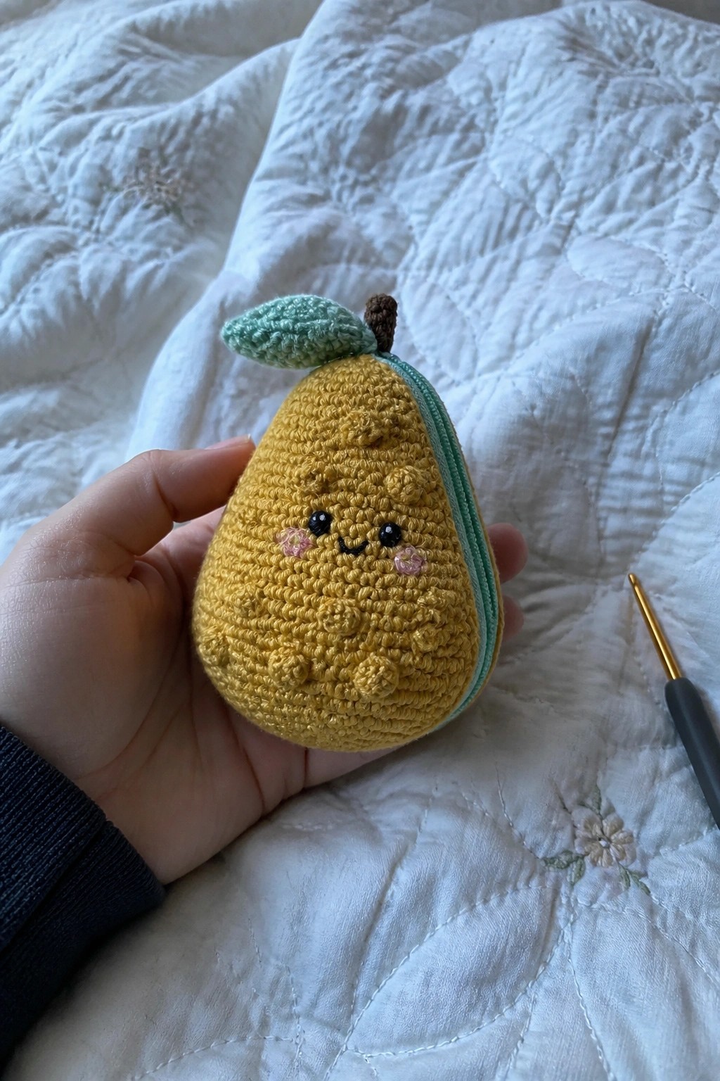 Hand holding yellow crocheted pear pouch with face on white quilt beside hook