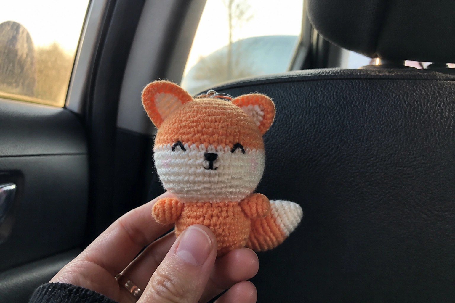 Hand holding small orange crocheted fox keychain inside a car near window.