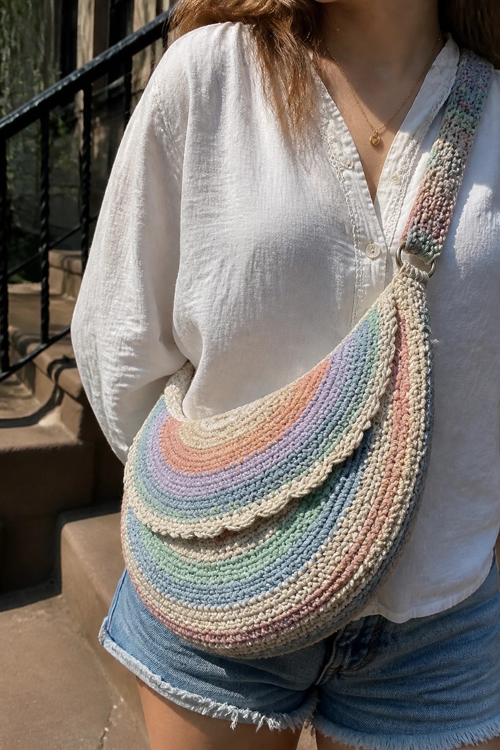 Woman in white shirt and denim shorts with pastel rainbow crocheted crescent bag.