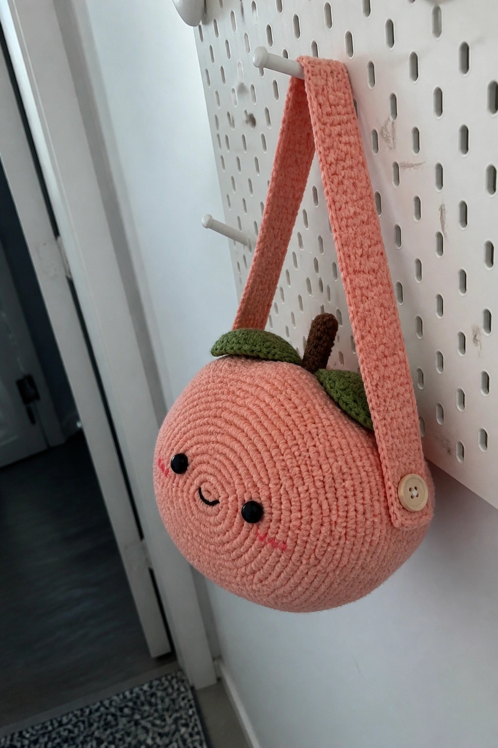 Crocheted peach bag with smiling face and strap hanging on white pegboard.