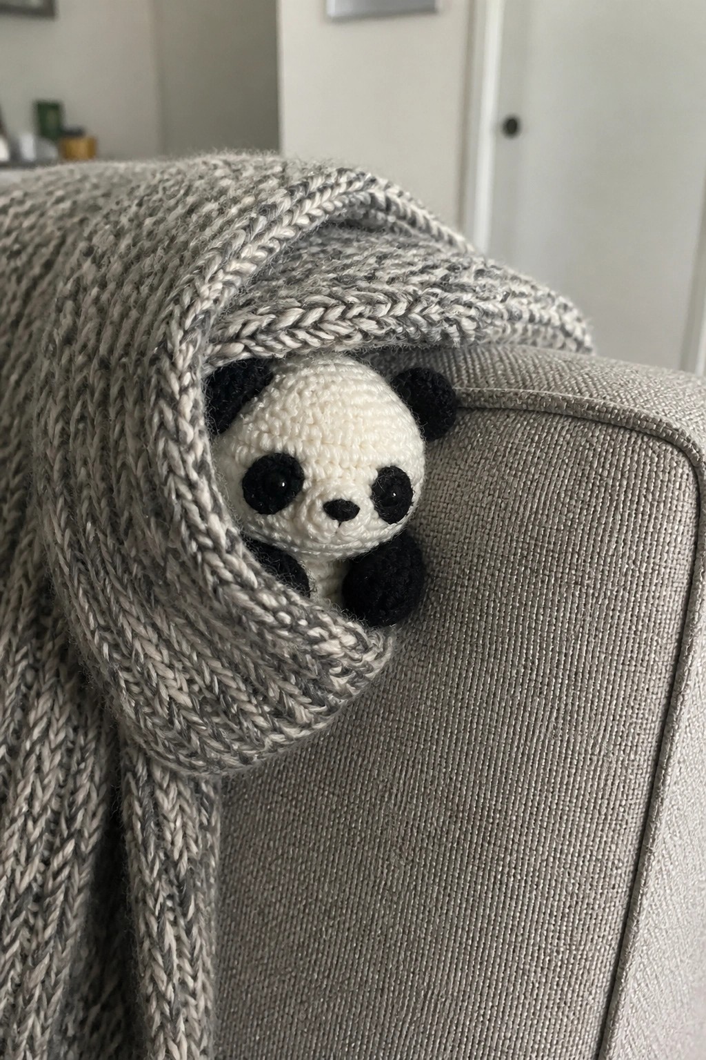 Crocheted panda peeking from under gray knitted blanket on gray couch.