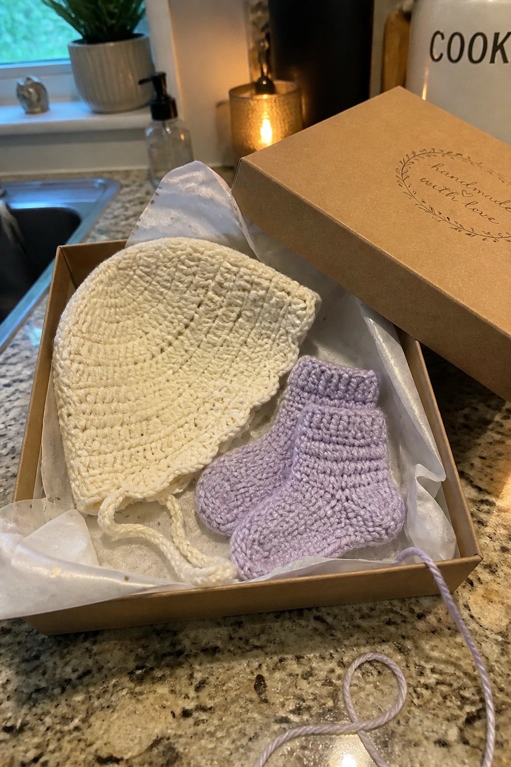 Open gift box with white crocheted baby bonnet and purple crocheted booties on tissue paper