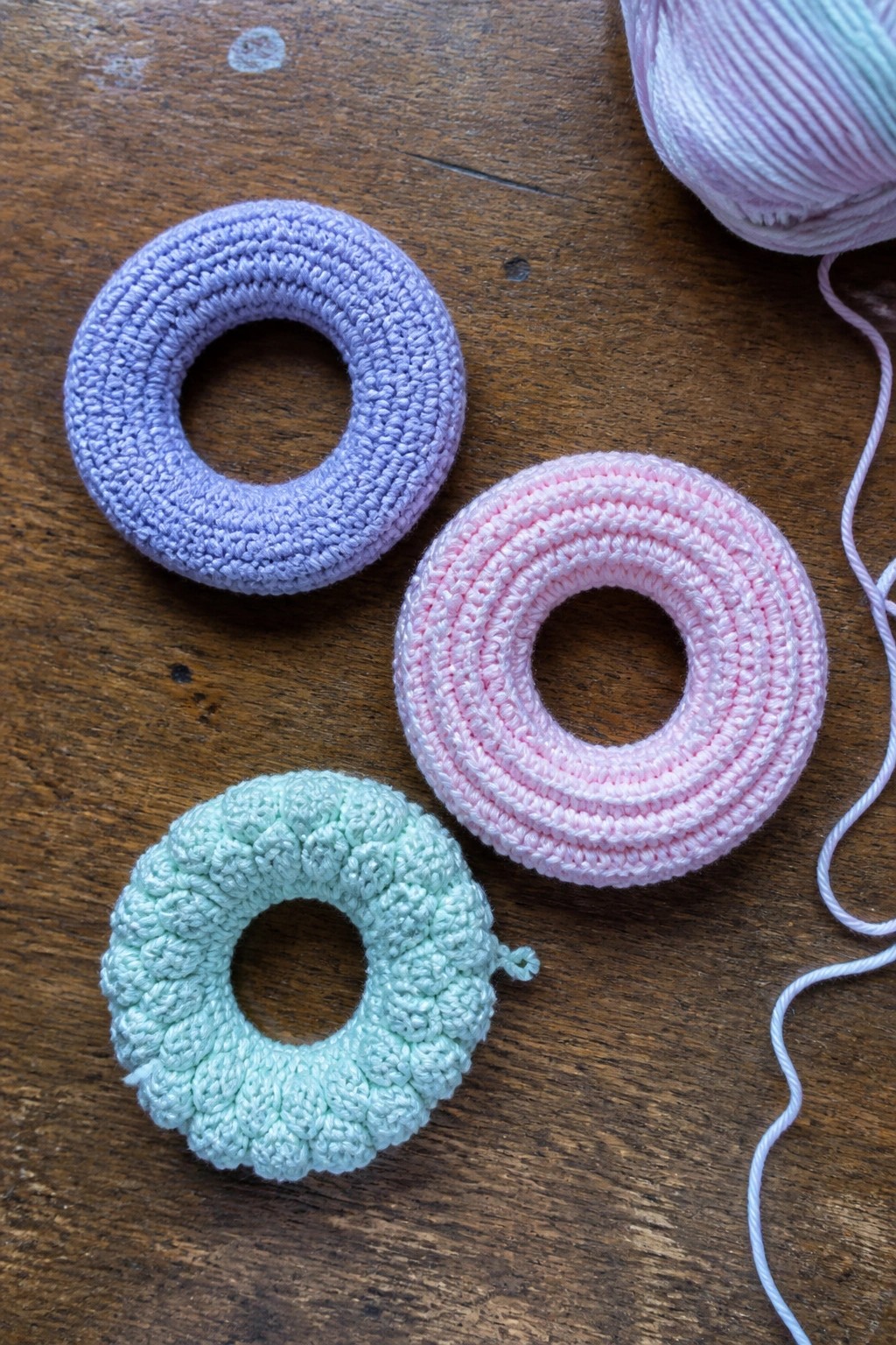 Three pastel crocheted rings in purple, pink, and mint on wood with yarn.