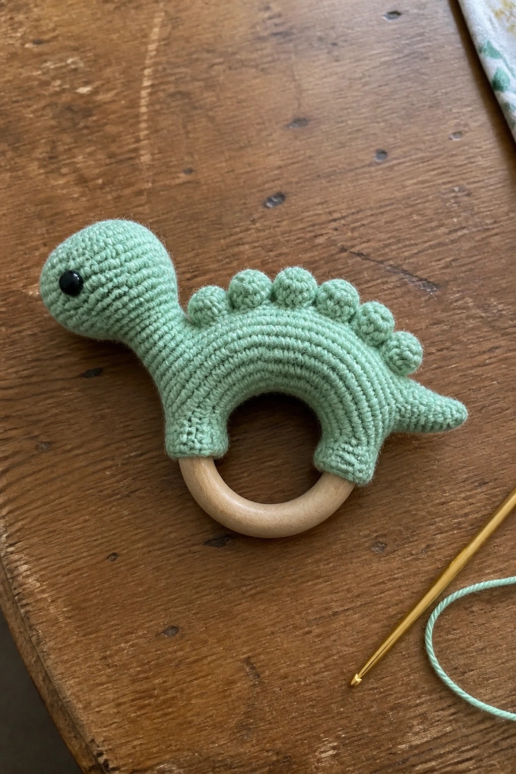Mint green crocheted dinosaur teether on wooden ring beside crochet hook