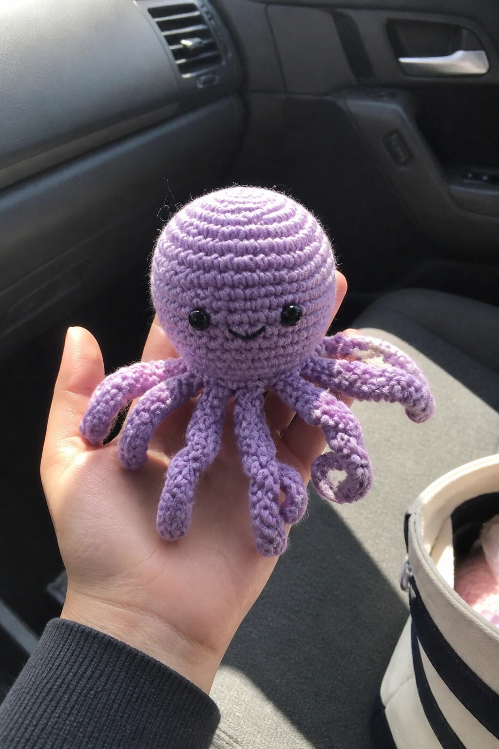 A small purple crocheted octopus plush with a smiling face held in a hand inside a car.