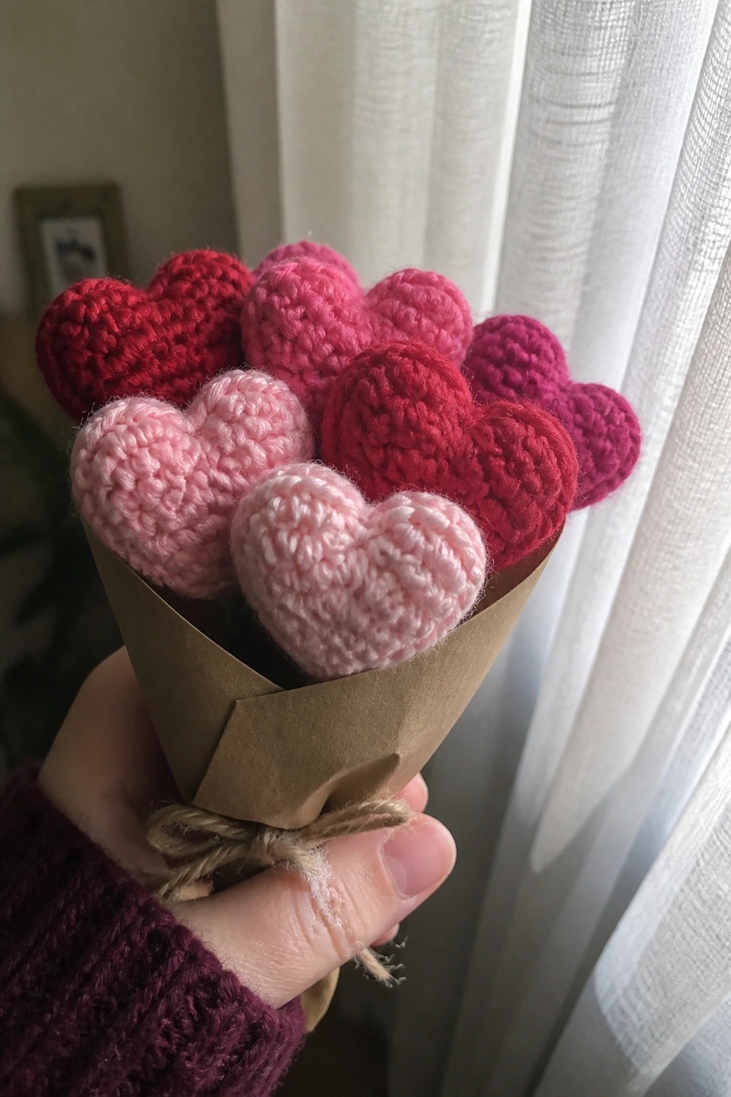Hand in purple sleeve holding crocheted pink-red hearts bouquet in brown paper.