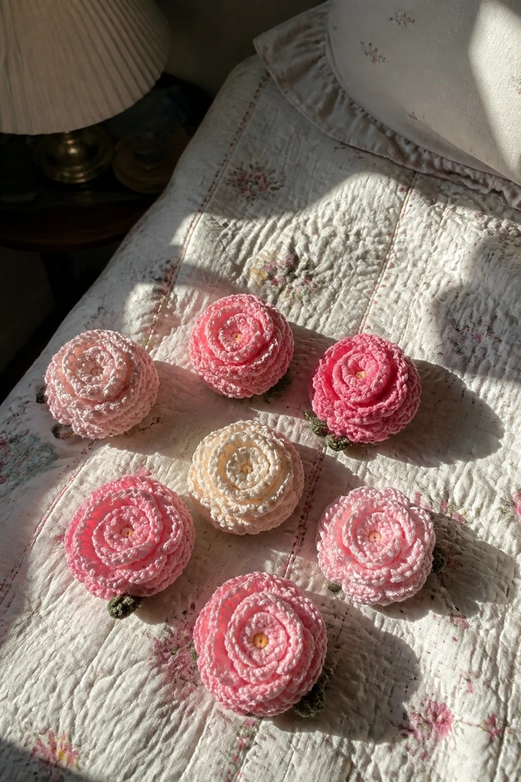 Crocheted pink and white roses arranged on a sunlit floral quilted bedspread.