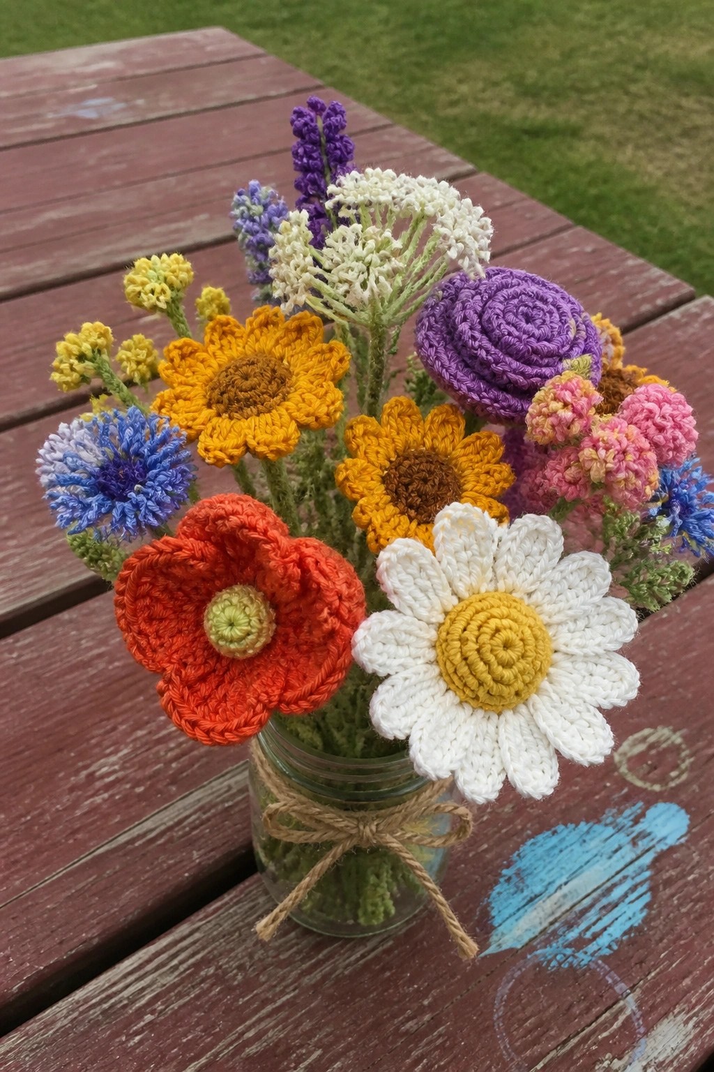 Colorful crocheted flowers in a glass jar on a wooden picnic table.