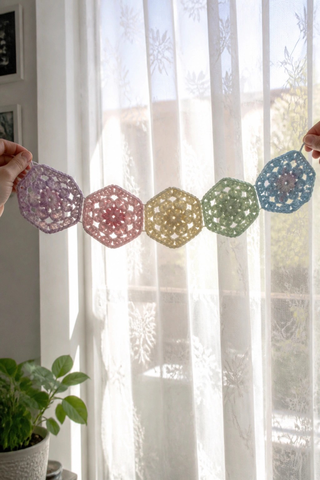 Hands holding pastel crocheted hexagon garland before sheer white curtains.