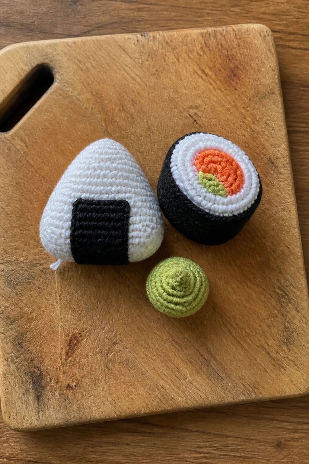 Crocheted sushi: white onigiri, nori maki roll with salmon, green wasabi on wood.