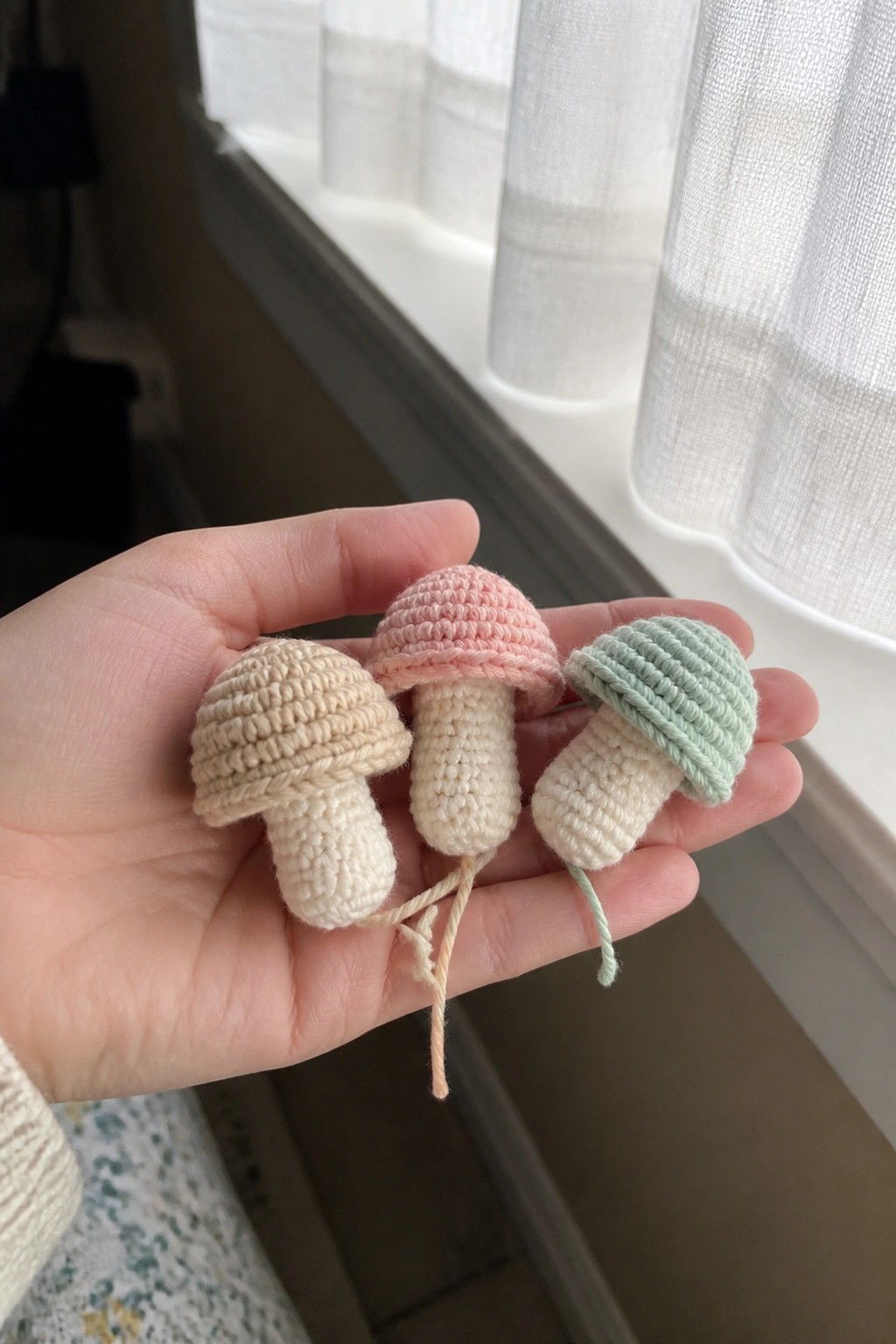 Hand holding three small crocheted mushrooms with beige, pink, and mint caps.