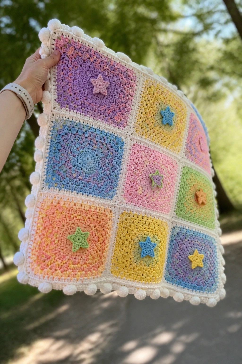 Hand holding pastel crochet blanket with star squares and white pom-pom border outdoors.