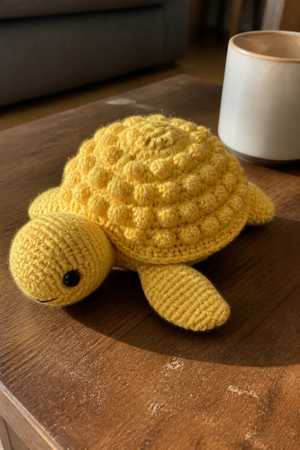 Yellow crocheted turtle on wooden table beside white ceramic mug.