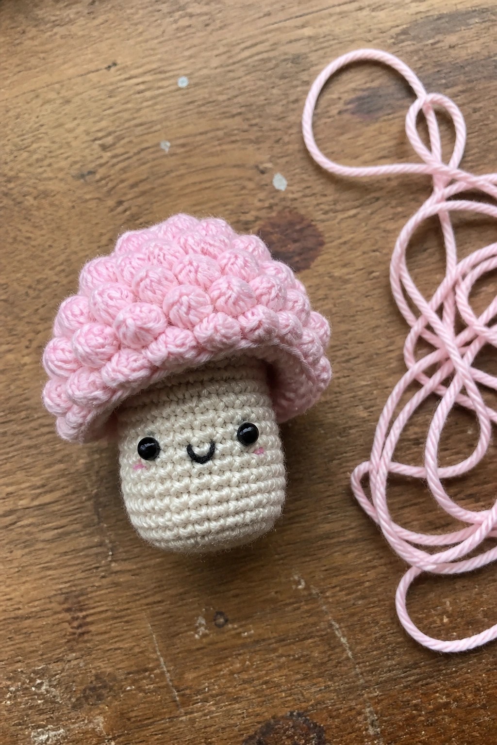 Pink crocheted mushroom toy with smiling face beside tangled pink yarn on wood