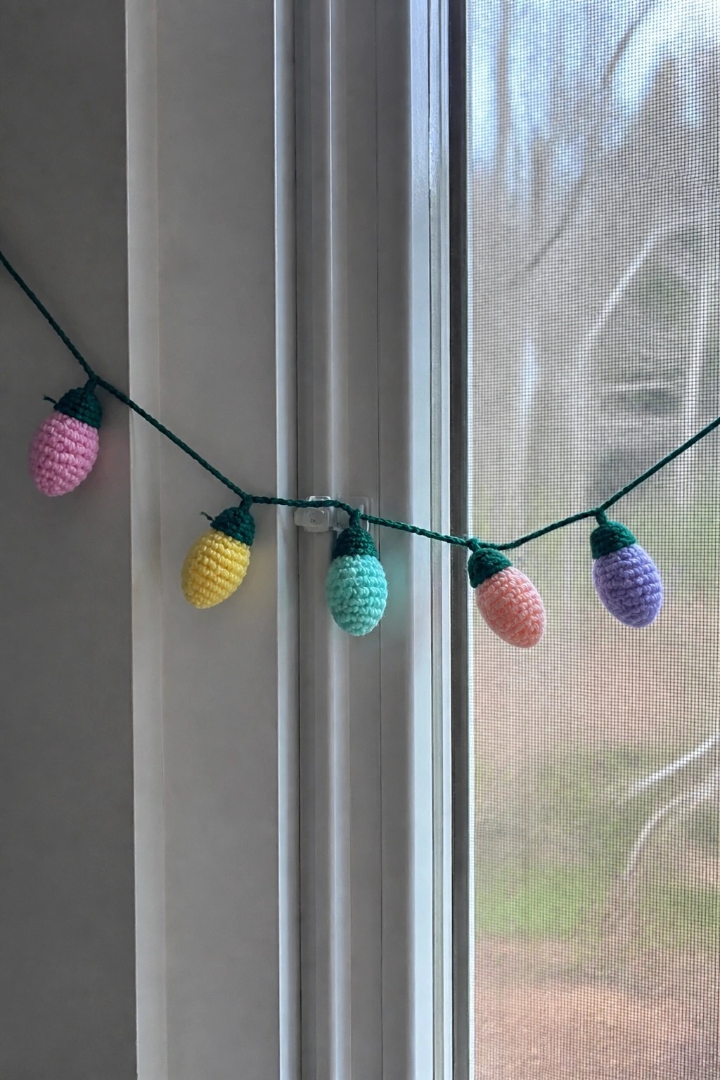 Crocheted pastel light bulb shapes in pink, yellow, mint, peach, and purple hang on a green yarn string across a window frame.