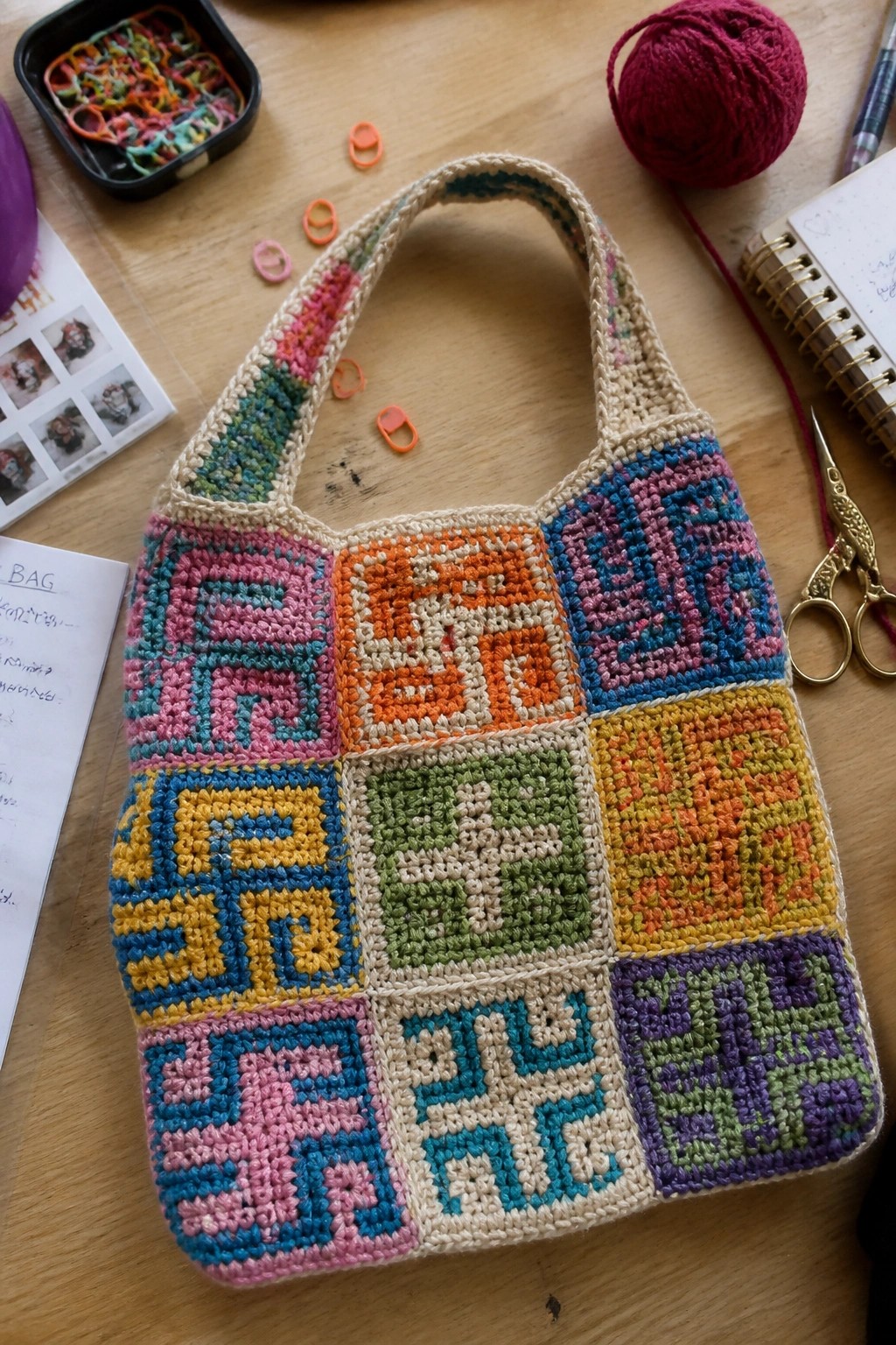 Crocheted sling bag assembled from colorful geometric granny squares on a wooden table with yarn and tools nearby.
