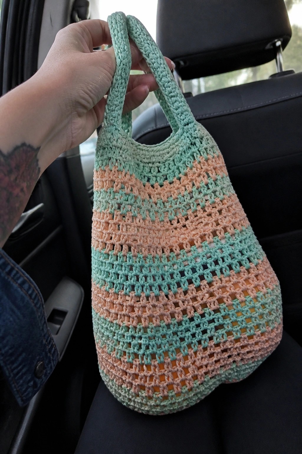Hand with rose tattoo holding striped mint-peach crocheted bag in car.