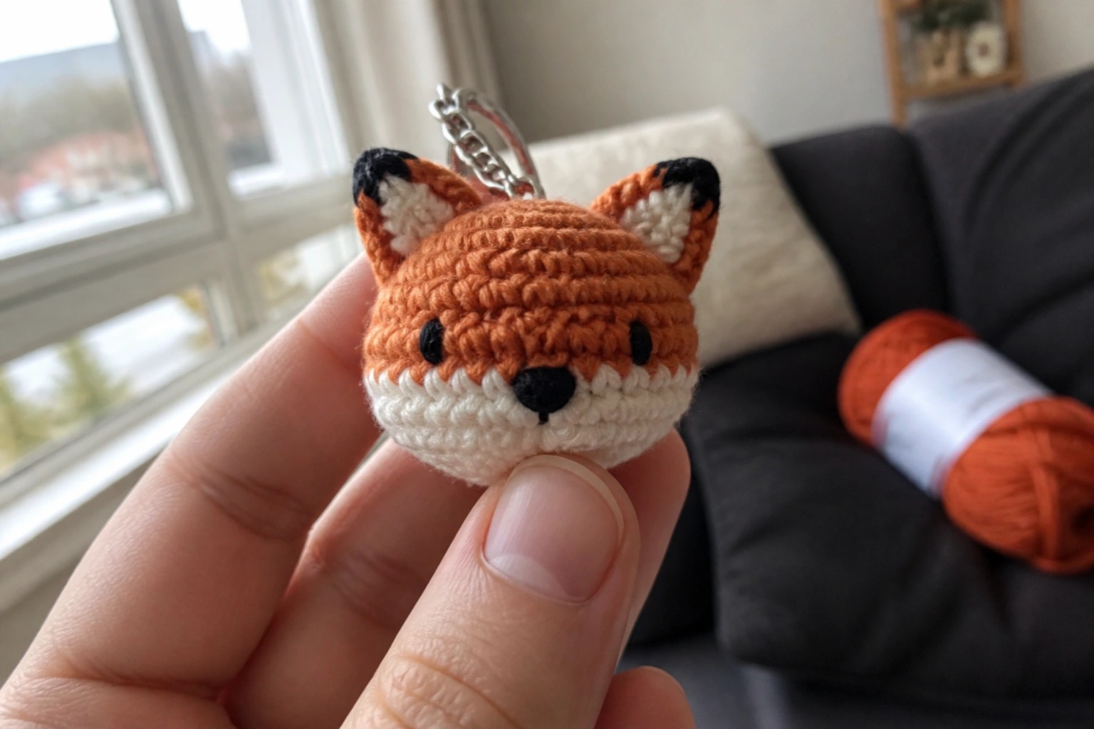 Hand holding crocheted orange fox keychain beside yarn ball and hook