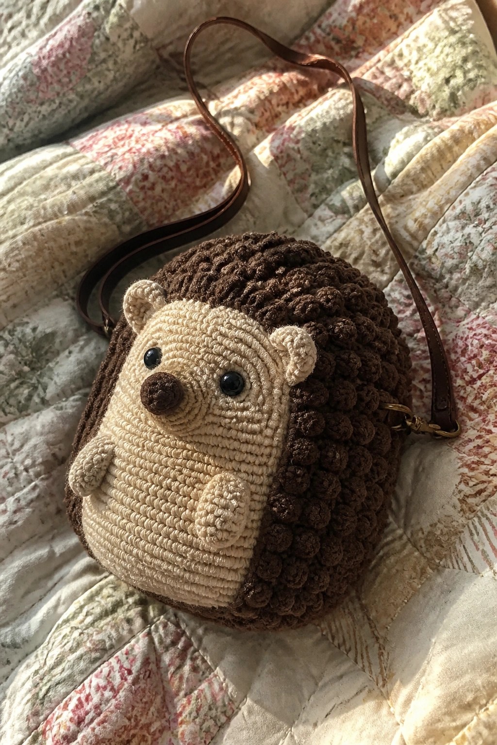 Crocheted hedgehog bag with beige face, brown spikes, and leather strap on quilt.