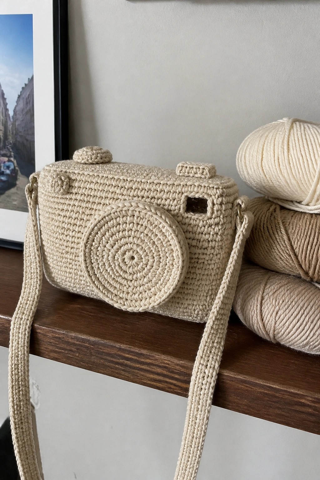 Crocheted beige camera-shaped bag with strap beside yarn balls on wood shelf