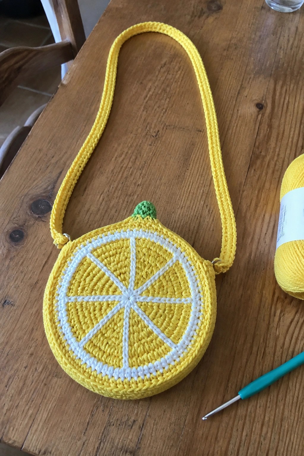 A yellow crocheted round bag resembling a lemon slice with white segments and a matching strap on a wooden table.