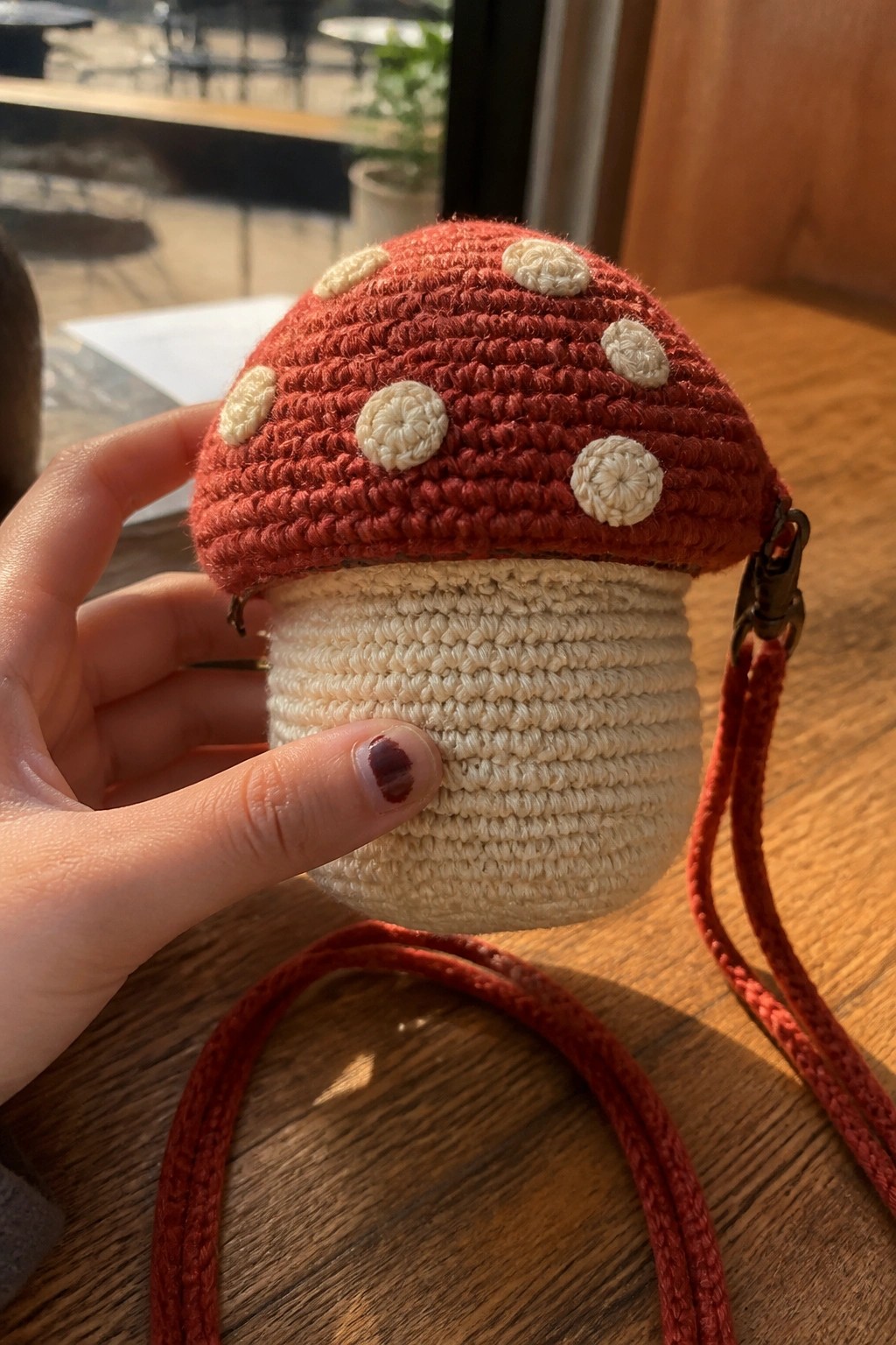 Hand holding crocheted mushroom pouch with red spotted cap, white base, and red strap.