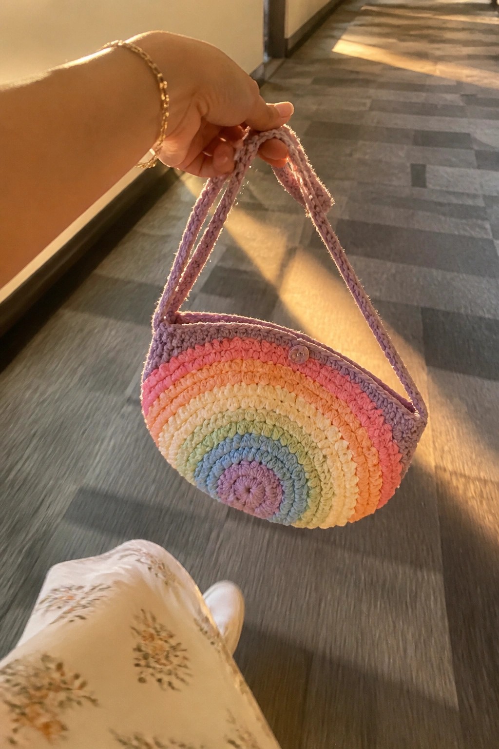 Hand holding rainbow crocheted crescent bag above floral skirt in sunny hallway