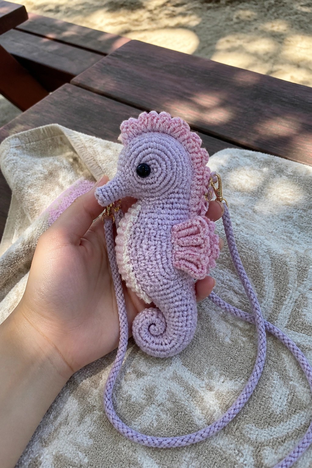 Hand holding pastel purple crocheted seahorse purse on towel over wooden bench.